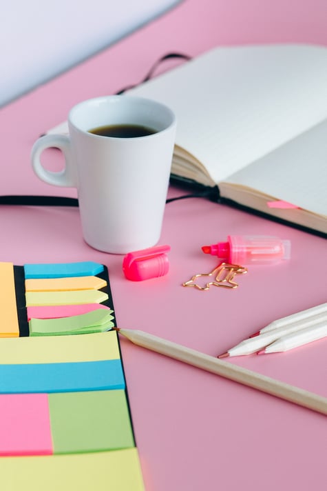 Colorful sticky notes near a white ceramic coffee mug. Mug is filled with coffee and near a blank open notebook. The items are on a pink backdrop. There are pink pencils, a pink highlighter, and a unicorn paperclip scattered between the notebook, coffee mug, and sticky notes.