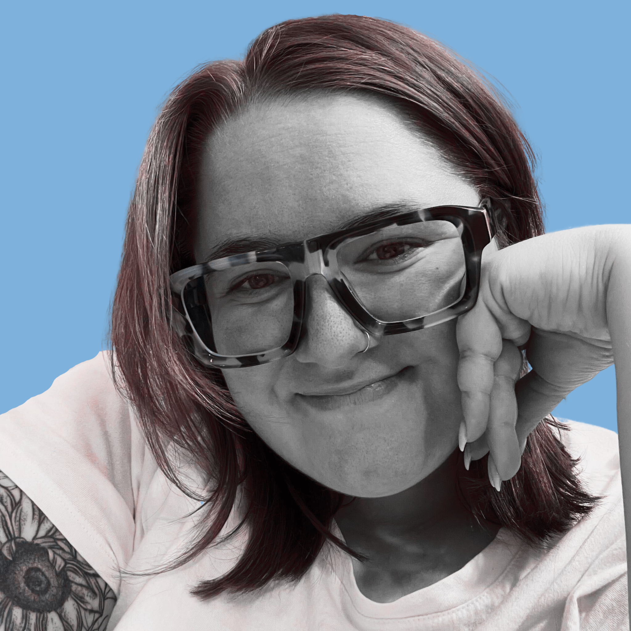 Emily is smiling with her head tilted to the reader's left, her left hand folded in half, resting on her left cheek. The photo is black and white with a reddish/pink tint. Emily is wearing a white t-shirt. A sunflower tattoo is near Emily's sleeve on the reader's left. The background is solid and a light baby blue. Emily is wearing black and white tortoiseshell, square glasses. 